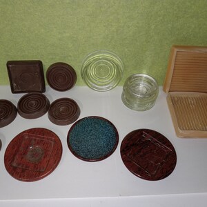 May include: An assortment of coasters and small decorative items. Includes brown, wooden-look coasters, clear glass coasters, and a coaster with a teal fabric insert. Some have textured surfaces.