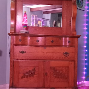 May include: A vintage wooden cabinet with a mirror, two drawers, and two doors. The cabinet has a carved leaf design on the doors and is finished in a rich brown color.