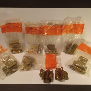 May include: A selection of brass-coloured cabinet hinges, some in their original plastic packaging with orange tape closures. The hinges are of various styles and sizes, suitable for different cabinetry projects.