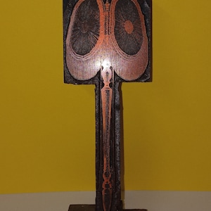 May include: A wooden printing block featuring a design of two stylised eyes with a stem and a base. The eyes are a reddish-brown colour and the stem and base are a darker brown.