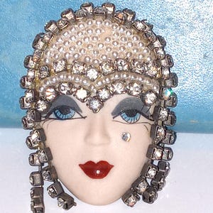 May include: A brooch featuring a stylized face with a pale complexion, blue eyes, and red lips. The figure wears a headpiece adorned with small white pearls and clear rhinestones, with additional rhinestone strands framing the face.