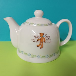 Puede incluir: Tetera de cerámica blanca con un diseño de oso de peluche. La tetera tiene un patrón floral azul y verde alrededor de la parte inferior y dice "Teddy Story" con el texto "Please eat I like something to sweet honey".