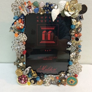 May include: A 5x7 inch black photo frame decorated with a variety of colorful jewels, beads, and trinkets. The frame is labeled "Copley Square" and "Halden International Designs".