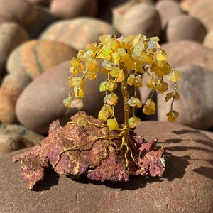 Peut inclure: Une sculpture artisanale d'arbre en fil de fer avec des feuilles de pierres précieuses jaunes. Le tronc et les branches de l'arbre sont en fil doré, et la base est en métal brun-rougeâtre texturé. L'arbre est exposé sur une grosse pierre brune arrondie.