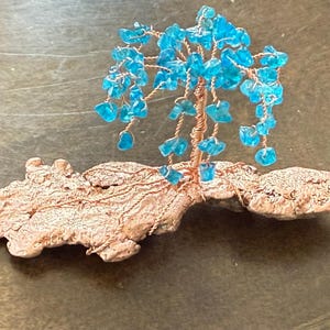 May include: A handcrafted wire tree sculpture with a copper trunk and branches, adorned with turquoise-colored glass beads. The tree sits on a textured, light brown base, creating a unique decorative piece.