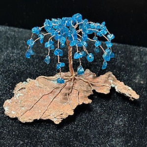 May include: A handcrafted wire tree sculpture with a copper trunk and branches, adorned with vibrant blue gemstone leaves. The tree is rooted in a textured, copper-coloured base, creating a miniature landscape. The sculpture is set against a dark background.