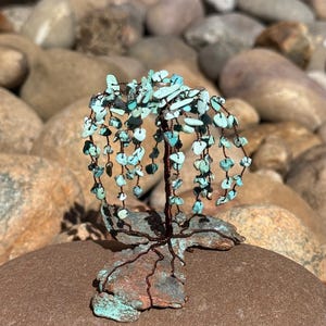 Könnte beinhalten: Eine handgefertigte Türkis- und Kupferdraht-Baumskulptur. Der Baum hat einen Kupferdrahtstamm und -äste, die mit kleinen Türkissteinen verziert sind. Der Sockel ist ein strukturiertes, kupferfarbenes Stück. Die Skulptur wird auf einem braunen Stein ausgestellt.