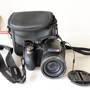 May include: A black Fujifilm FinePix digital camera with a Super EBC Fujinon lens featuring 24x zoom, 4.3-103.2mm focal length, and 1:3.1-5.9 aperture. The camera includes a black neck strap with the Fujifilm logo and a black carrying case.