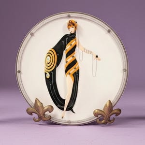 May include: A round white decorative plate featuring a stylized illustration of a figure in a black and orange striped dress with a long flowing wrap. The plate has a silver-toned rim and is supported by two gold-colored metal fleur-de-lis stands.