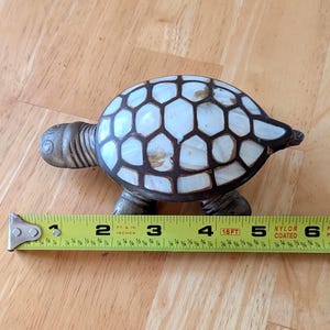 May include: A turtle-shaped decorative object with a metallic head and legs and a shell featuring white, iridescent, hexagonal panels outlined in dark metal. A yellow measuring tape placed alongside shows the object is approximately 6 inches long.