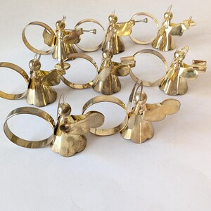 May include: A set of eight polished brass napkin rings, each featuring a stylized angel figure with wings and a halo. The angels are depicted in various poses, some holding small instruments or objects, attached to circular brass bands.