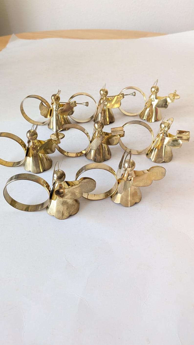 May include: A set of eight polished brass napkin rings, each featuring a stylized angel figure with wings and a halo. The angels are depicted in various poses, some holding small instruments or objects, attached to circular brass bands.
