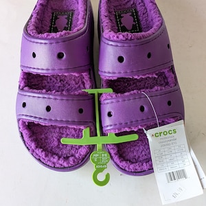May include: A pair of purple Crocs Cozzzy sandals featuring two wide straps with ventilation holes and a plush, fuzzy purple interior lining. A green plastic hanger connects the sandals, and a white product tag displays sizing information.