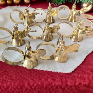 May include: A set of eight gold-toned metal napkin rings, each featuring a stylized angel figure. The angels are depicted with wings and various poses, including some holding small trumpets or books, attached to circular metal bands.