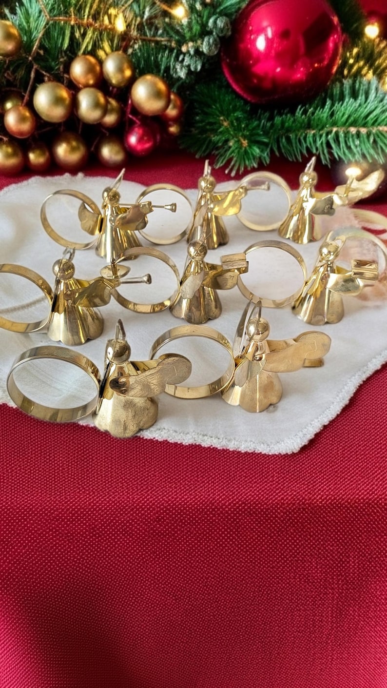 May include: A set of eight gold-toned metal napkin rings, each featuring a stylized angel figure. The angels are depicted with wings and various poses, including some holding small trumpets or books, attached to circular metal bands.