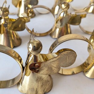 May include: A collection of polished brass napkin rings, each featuring a stylized angel figure with a circular halo and wings. The metallic gold-toned ornaments are arranged in a group, showcasing a smooth, reflective surface and a conical base.