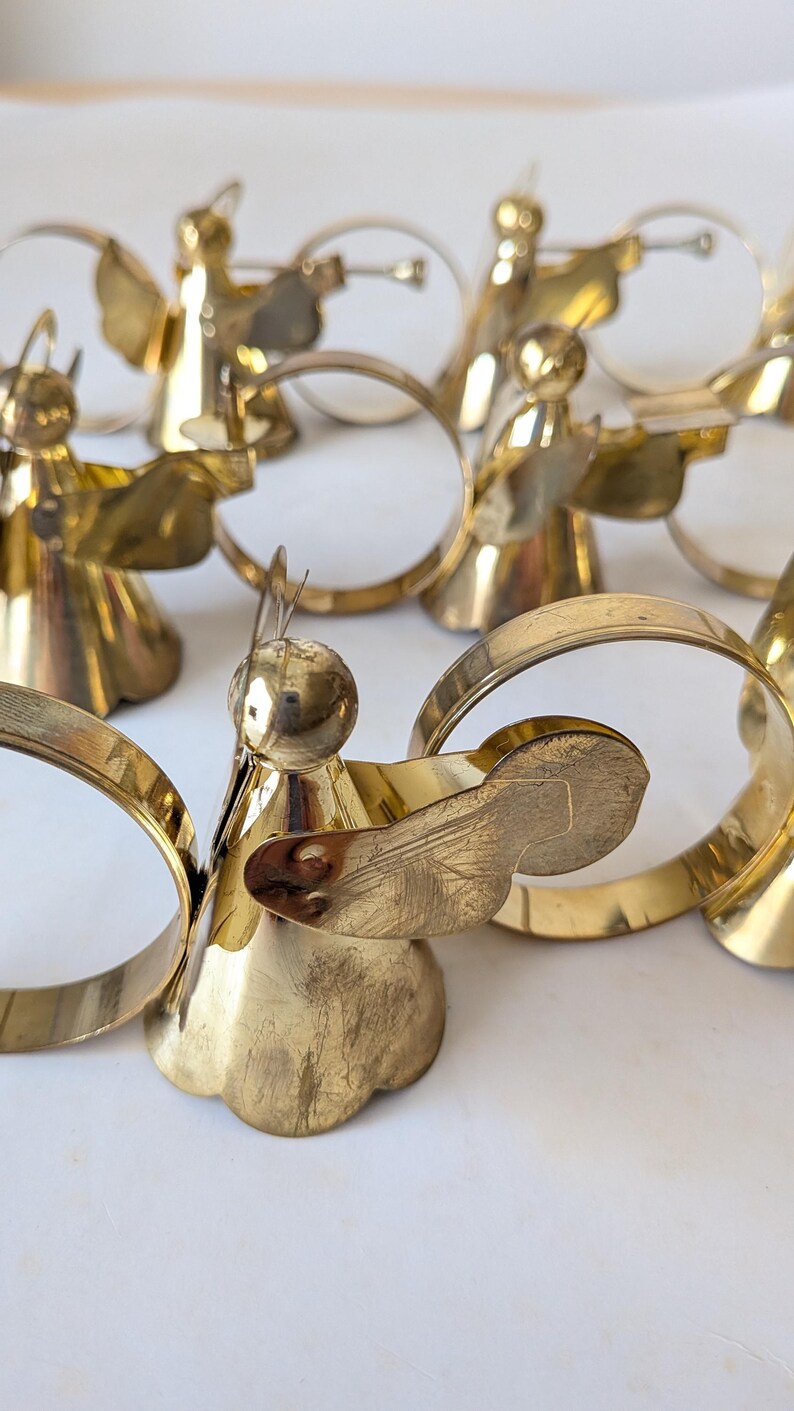 May include: A collection of polished brass napkin rings, each featuring a stylized angel figure with a circular halo and wings. The metallic gold-toned ornaments are arranged in a group, showcasing a smooth, reflective surface and a conical base.