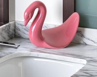 May include: A glossy, solid pink ceramic swan-shaped dish with a smooth, curved body and an elongated neck. The swan is depicted in a resting position with its head turned slightly downward, featuring a minimalist design and a reflective finish.