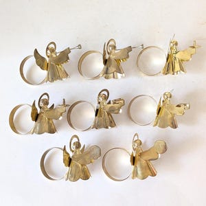 May include: A set of eight gold-toned metal napkin rings, each featuring a stylized angel figure with a halo and wings. The angels are depicted in various poses, some holding small instruments or objects, attached to a circular metal band.