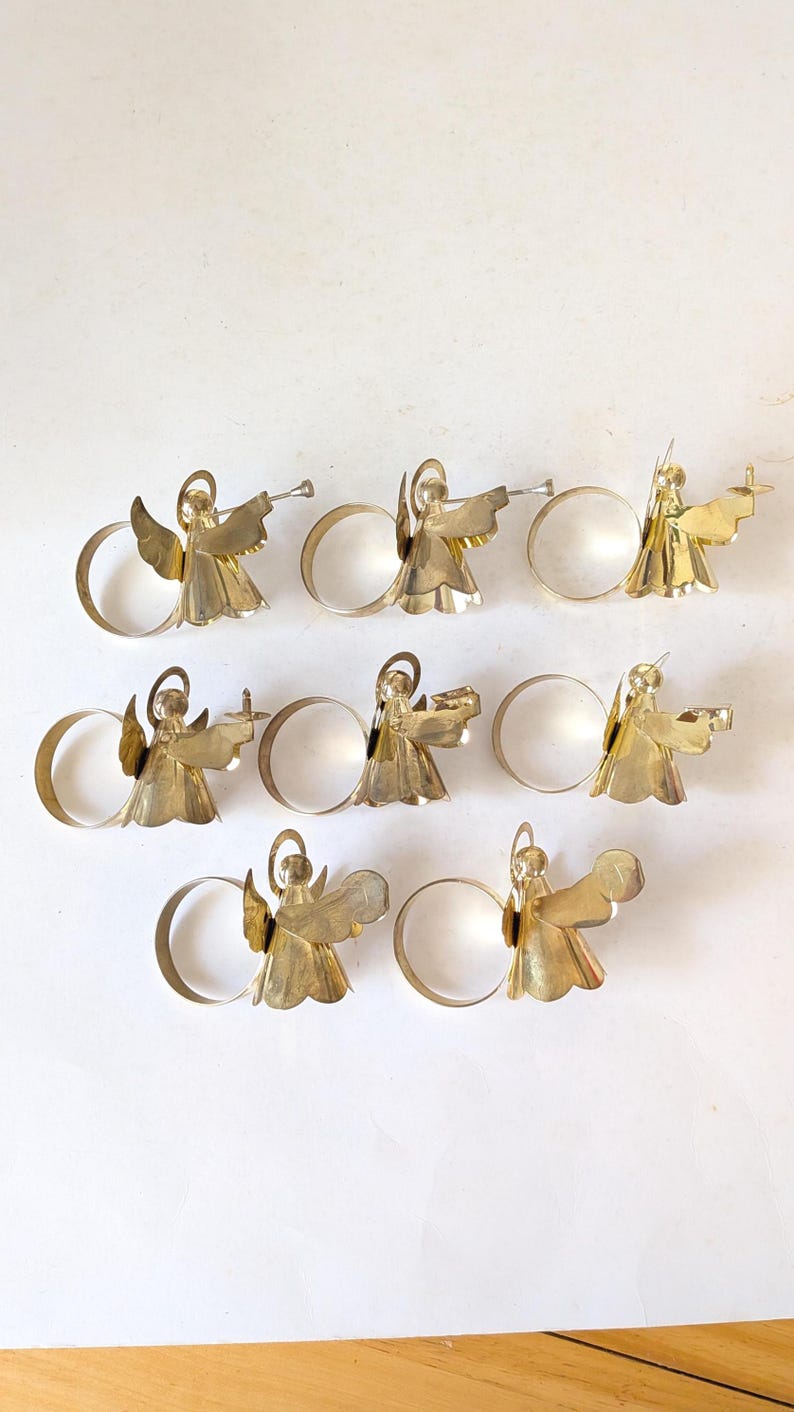 May include: A set of eight gold-toned metal napkin rings, each featuring a stylized angel figure with a halo and wings. The angels are depicted in various poses, some holding small instruments or objects, attached to a circular metal band.