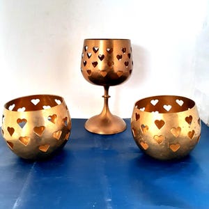 May include: Three copper-colored candle holders. Two are round with heart-shaped cutouts, and one is a stemmed goblet with similar heart cutouts. The objects are on a blue surface.