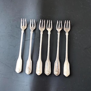 May include: A set of six silver-toned metal cocktail forks arranged in a row. Each fork features a three-pronged head and a slender handle that terminates in a decorative shell motif at the base. The metal has a polished, reflective finish.