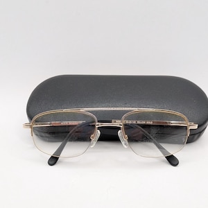 May include: Gold-tone metal eyeglasses with a double bridge and clear lenses. The inner frame is printed with M'EYE GENERATION, ROBIT 53-20-145 YELLOW GOLD. The glasses are positioned in front of a black, oval-shaped, hard-shell protective case.
