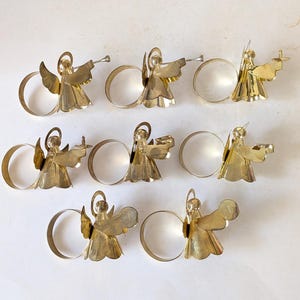 May include: A set of seven gold-toned metal napkin rings, each featuring a stylized angel figure with a halo and wings. The angels are depicted in various poses, some holding small objects like trumpets or candles, attached to a circular band.