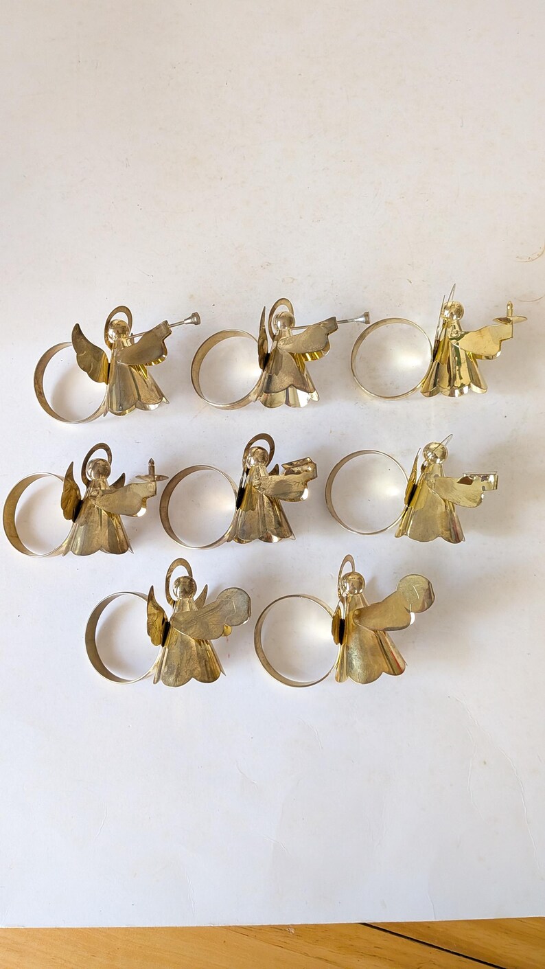 May include: A set of seven gold-toned metal napkin rings, each featuring a stylized angel figure with a halo and wings. The angels are depicted in various poses, some holding small objects like trumpets or candles, attached to a circular band.