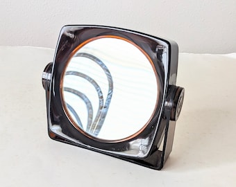 May include: A square-shaped vanity mirror with a dark brown, translucent acrylic frame. The circular mirror is mounted on a rotating axis, allowing it to tilt within the square housing. The design features smooth, rounded edges and a glossy finish.