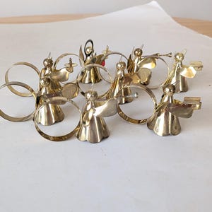 May include: A collection of eight polished brass napkin rings, each featuring a small angel figure. The angels are depicted in various poses, some holding instruments or books, with circular brass bands attached to their backs for holding napkins.