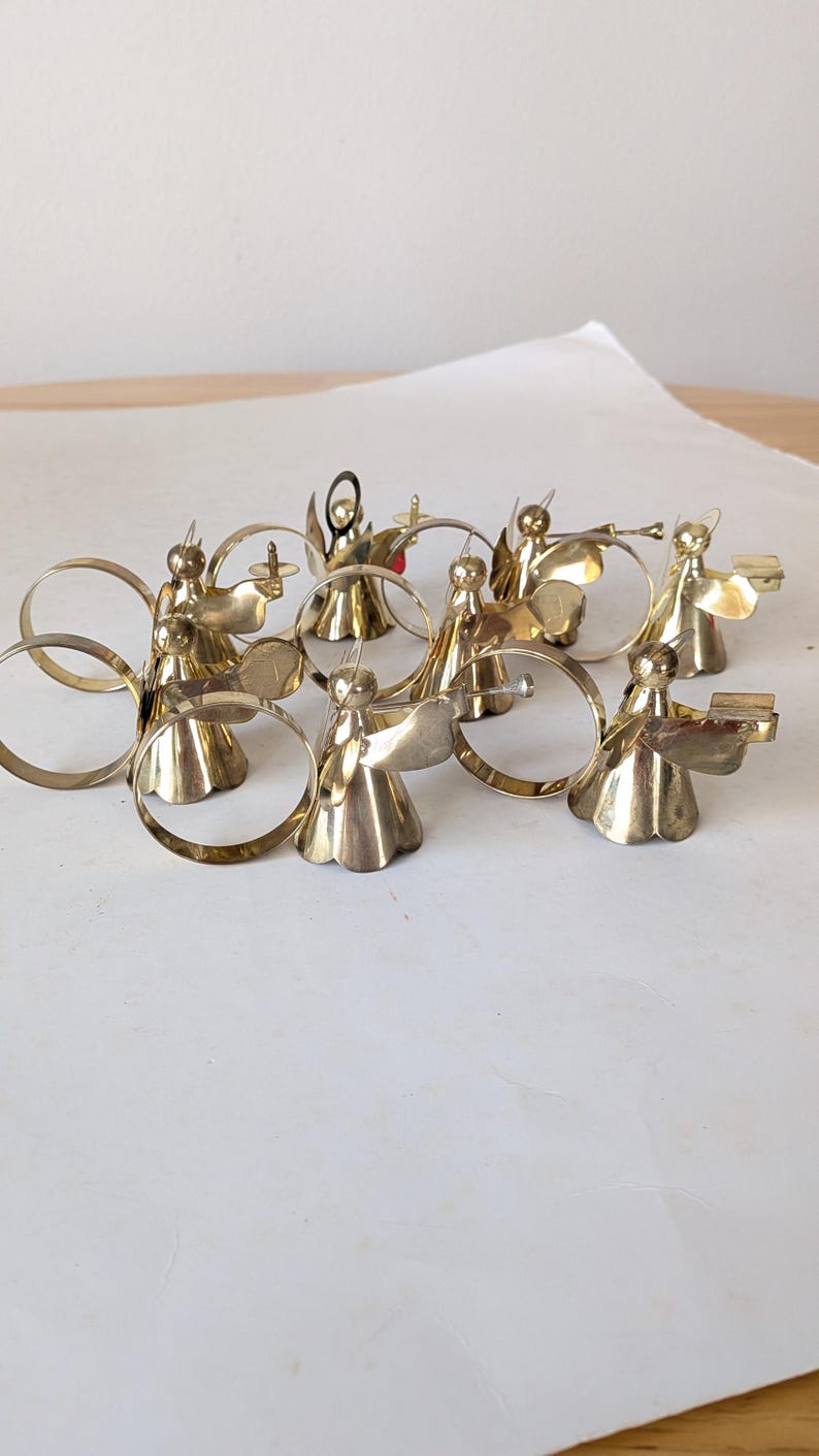 May include: A collection of eight polished brass napkin rings, each featuring a small angel figure. The angels are depicted in various poses, some holding instruments or books, with circular brass bands attached to their backs for holding napkins.