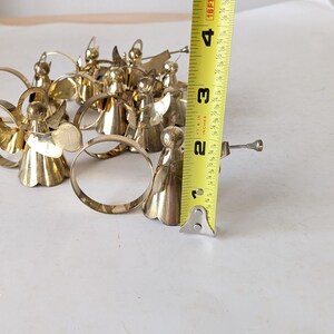 May include: A collection of gold-toned metal angel napkin rings, each featuring a small trumpet. A yellow measuring tape is positioned vertically next to the figures, indicating a height of approximately 2.5 inches for each angel ornament.