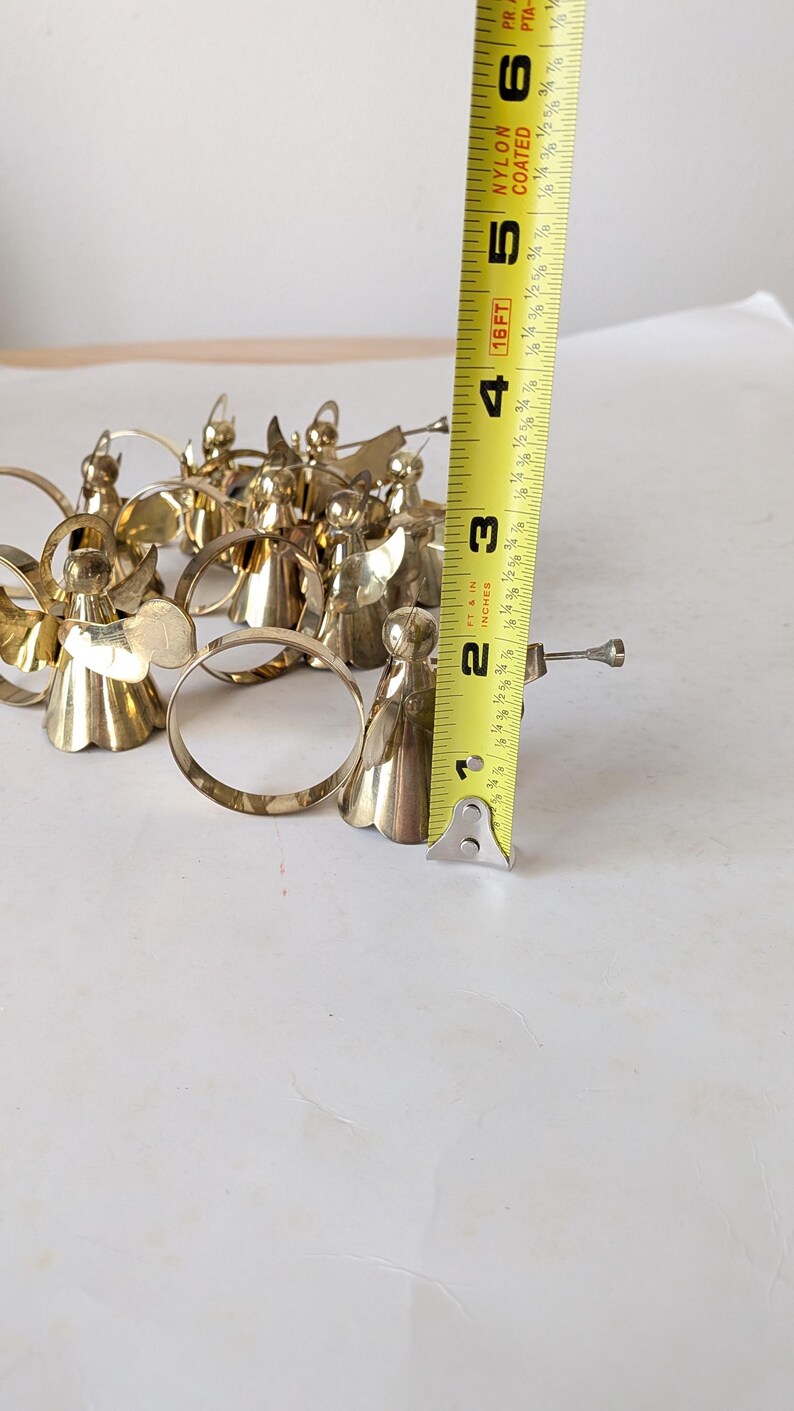 May include: A collection of gold-toned metal angel napkin rings, each featuring a small trumpet. A yellow measuring tape is positioned vertically next to the figures, indicating a height of approximately 2.5 inches for each angel ornament.