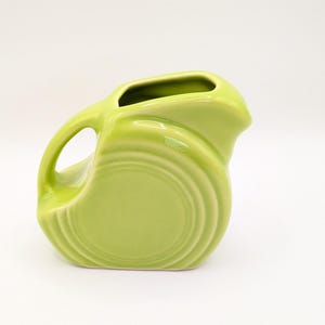 May include: A lime green ceramic pitcher with a rounded body and a handle. The pitcher has a spout and a circular design on the front. The handle is a loop shape. The pitcher is on a white background.