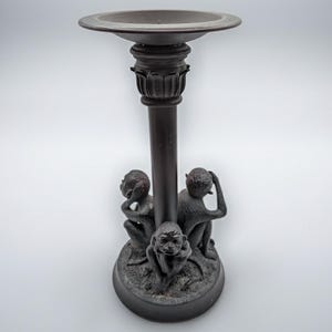 May include: Dark brown decorative candle holder with three monkey figurines at the base. The candle holder has a round top and a detailed column. The monkeys are in the "see no evil, hear no evil, speak no evil" poses.