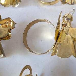 May include: A collection of gold-toned metal napkin rings, each featuring a stylized angel figure with a halo and outstretched wings. The angel figures are attached to a circular band, creating a decorative set for table settings.