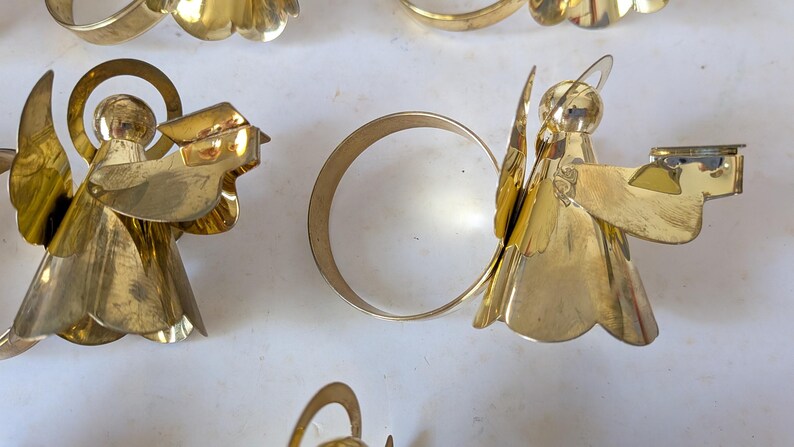 May include: A collection of gold-toned metal napkin rings, each featuring a stylized angel figure with a halo and outstretched wings. The angel figures are attached to a circular band, creating a decorative set for table settings.