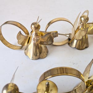 May include: A set of gold-colored metal napkin rings, each featuring a stylized angel figure with outstretched wings and a halo. The angel figures are attached to circular metal bands, creating a cohesive design for table settings.