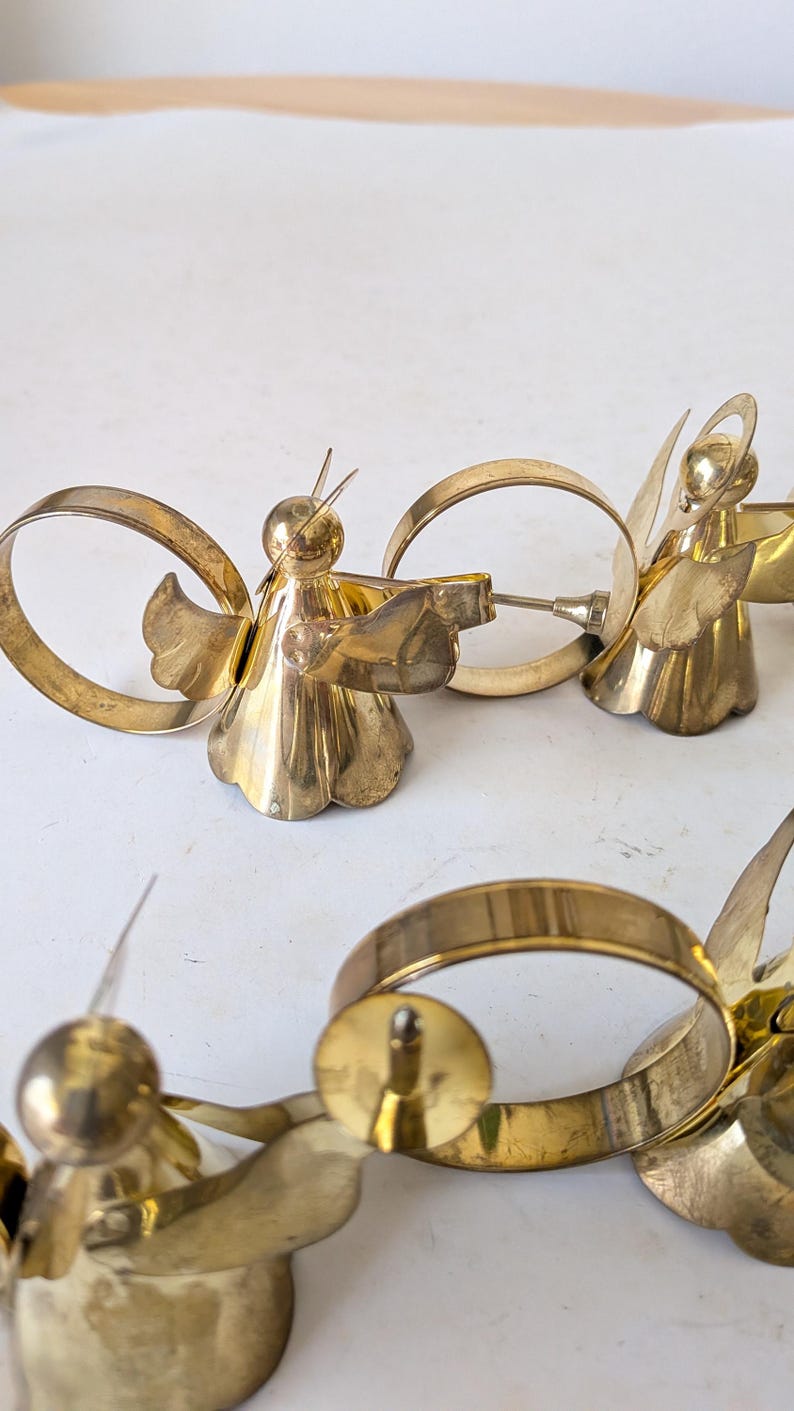 May include: A set of gold-colored metal napkin rings, each featuring a stylized angel figure with outstretched wings and a halo. The angel figures are attached to circular metal bands, creating a cohesive design for table settings.