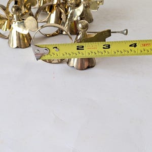 May include: A collection of gold-toned metal angel figurines, each featuring a circular halo and a trumpet. A yellow measuring tape is positioned across the front, showing the circular base of one angel measures approximately 2 inches in diameter.