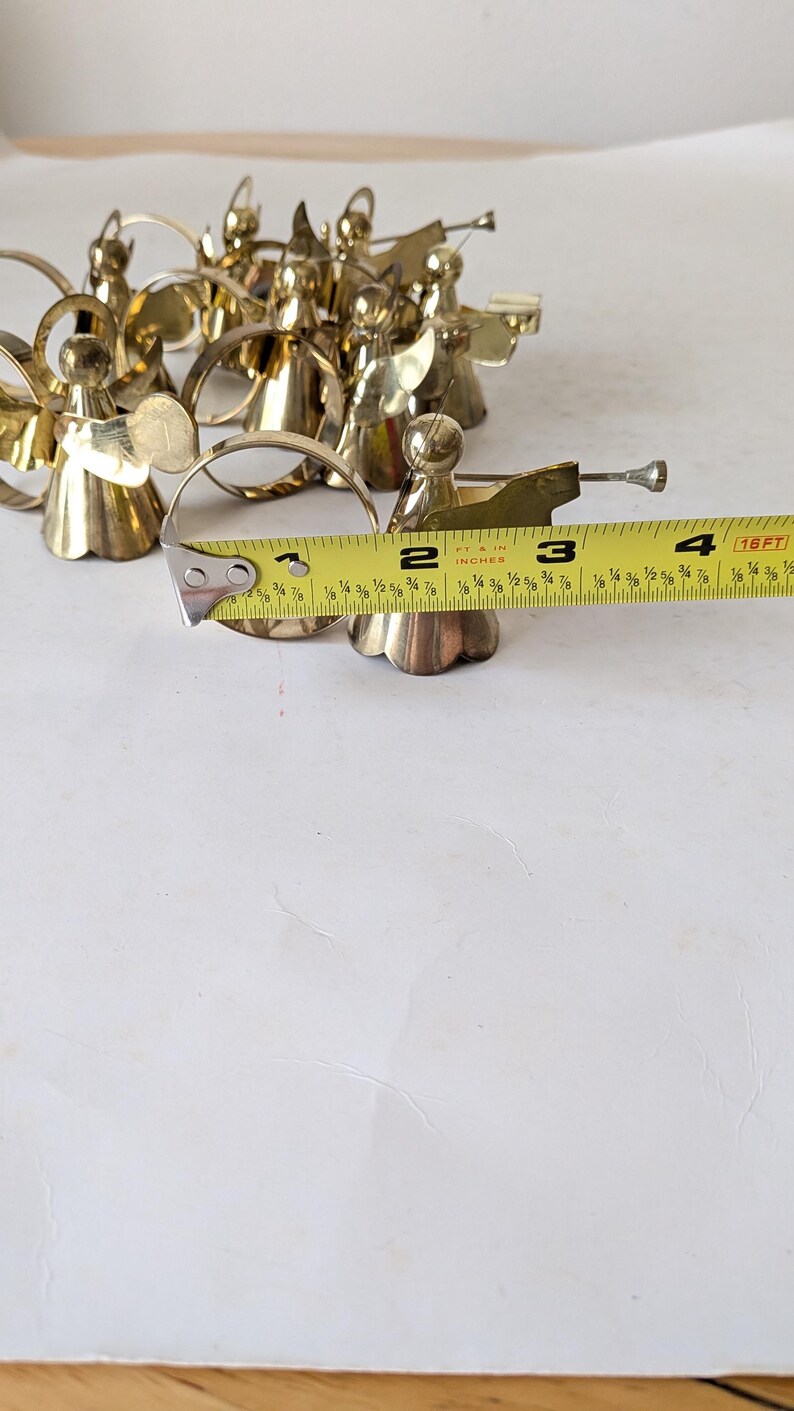 May include: A collection of gold-toned metal angel figurines, each featuring a circular halo and a trumpet. A yellow measuring tape is positioned across the front, showing the circular base of one angel measures approximately 2 inches in diameter.