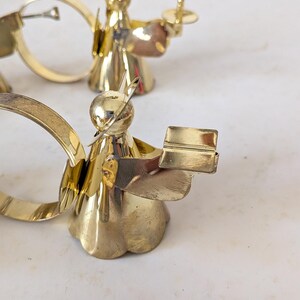May include: Two polished brass napkin rings shaped like angels. Each angel figure features a smooth, rounded head with a thin wire halo, a flared skirt base, and outstretched arms holding a small, rectangular book-shaped object in front.