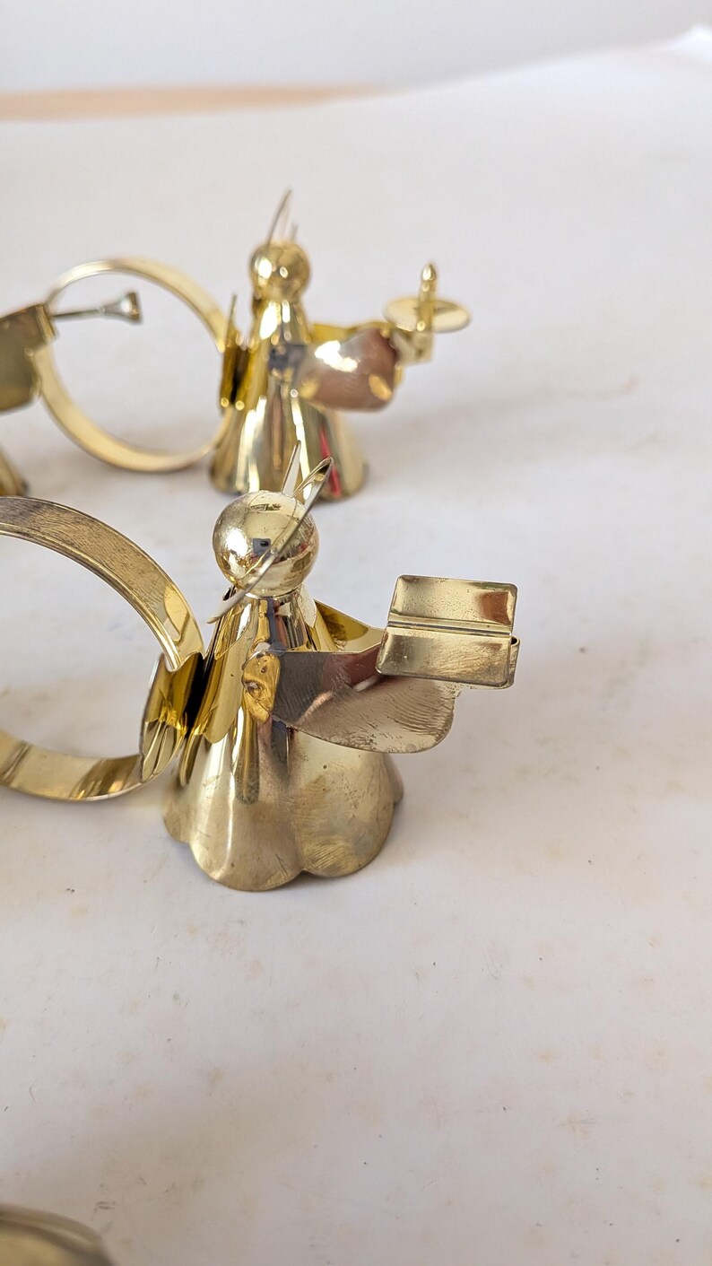 May include: Two polished brass napkin rings shaped like angels. Each angel figure features a smooth, rounded head with a thin wire halo, a flared skirt base, and outstretched arms holding a small, rectangular book-shaped object in front.