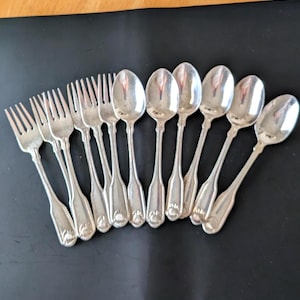 May include: A set of eleven silver-toned metal utensils consisting of three forks and eight spoons. Each piece features a slender handle with a decorative shell-like motif at the base. The utensils are arranged in a fan-like pattern on a dark surface.