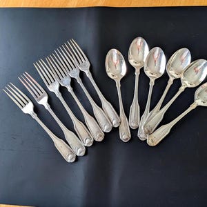 May include: A set of silver-toned metal flatware consisting of four forks and five spoons. Each piece features a polished finish and a decorative shell motif embossed at the base of the handle. The utensils are arranged in a fan-like pattern.
