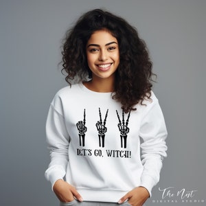 May include: White sweatshirt with a black skeleton hand graphic and the text "Let's go, witch!"
