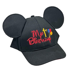 May include: A black baseball cap featuring two large circular ears on top. The front is embroidered with the red text My 1st Birthday, with the number 1 in yellow and a small cluster of red, blue, and yellow balloons attached to the text.