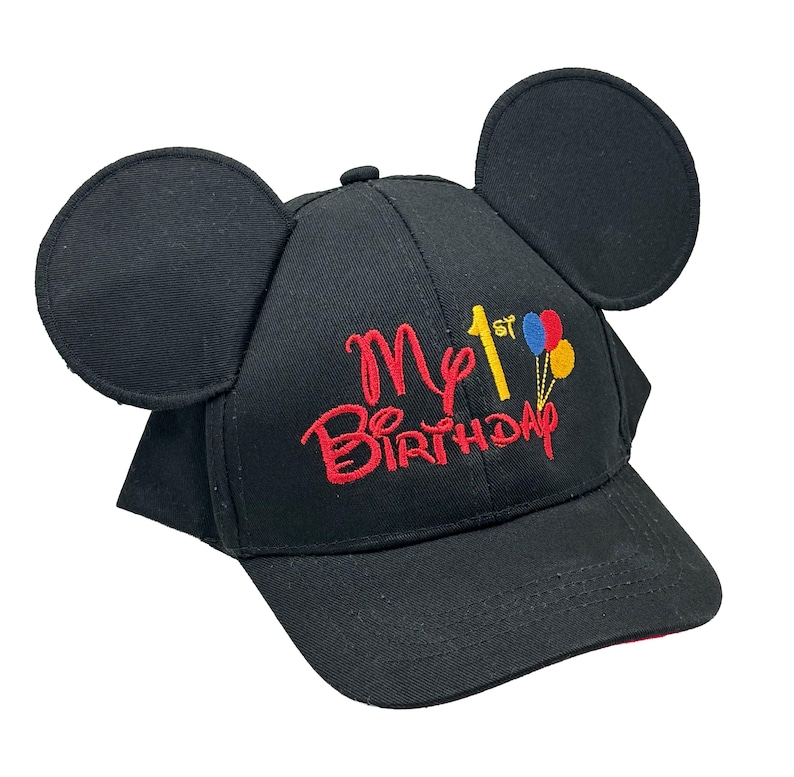 May include: A black baseball cap featuring two large circular ears on top. The front is embroidered with the red text My 1st Birthday, with the number 1 in yellow and a small cluster of red, blue, and yellow balloons attached to the text.
