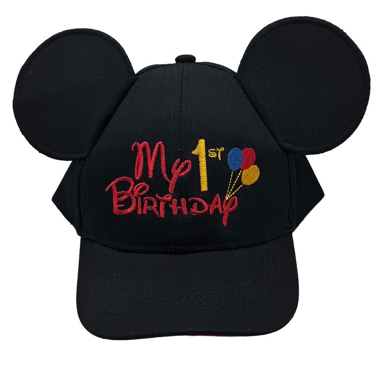 May include: A black baseball cap featuring two large circular ears on top. The front is embroidered with the red text "My 1st Birthday" and a small cluster of three balloons in blue, red, and yellow. The cap has a curved brim and a structured crown.